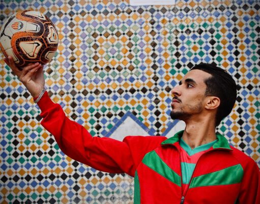 Achraf Hakimi : l’icône mode des Lions de l’Atlas Achraf Hakimi en costume de footballeur marocain posant devant un fond rouge